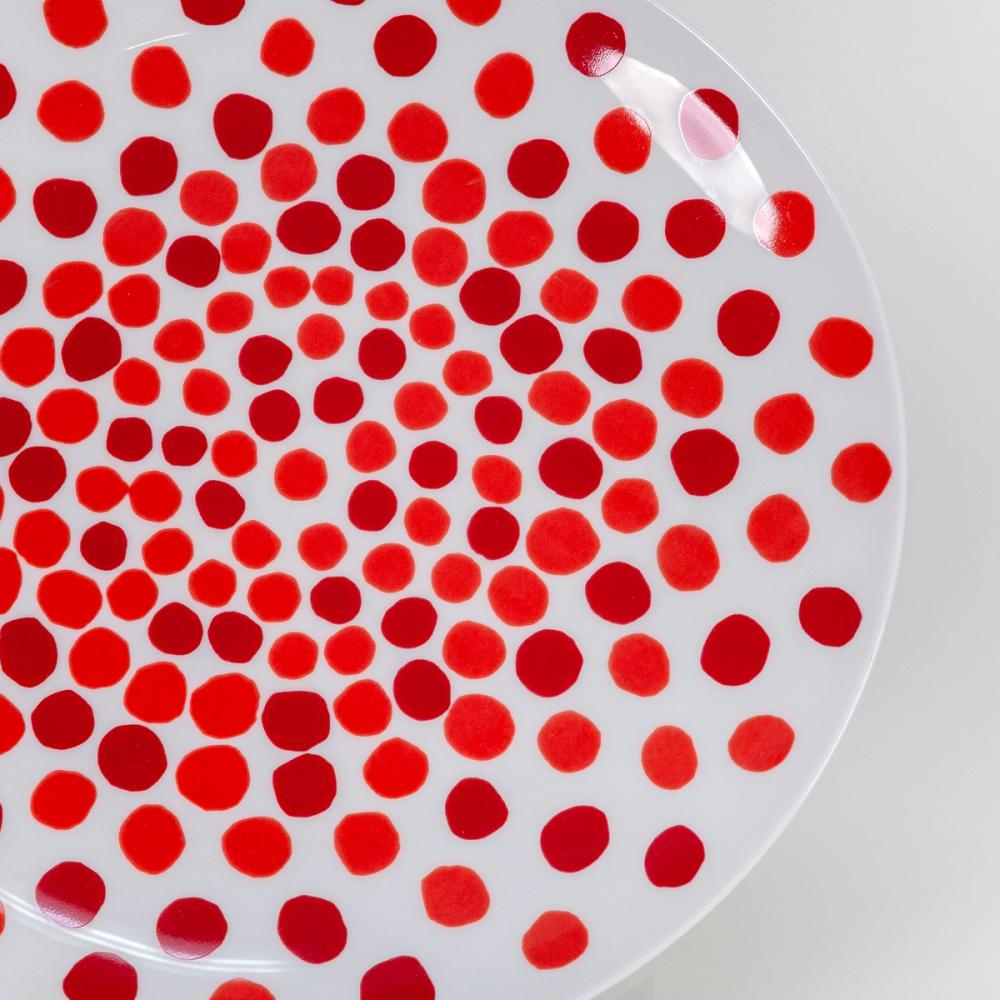 A detail photograph of the right edge to the center of a white plate with bold wobbly red dots in wobbly concentric circles. The dots are closer together in the center of the plate and become more spread out towards the edge. The dots do not vary greatly in size.