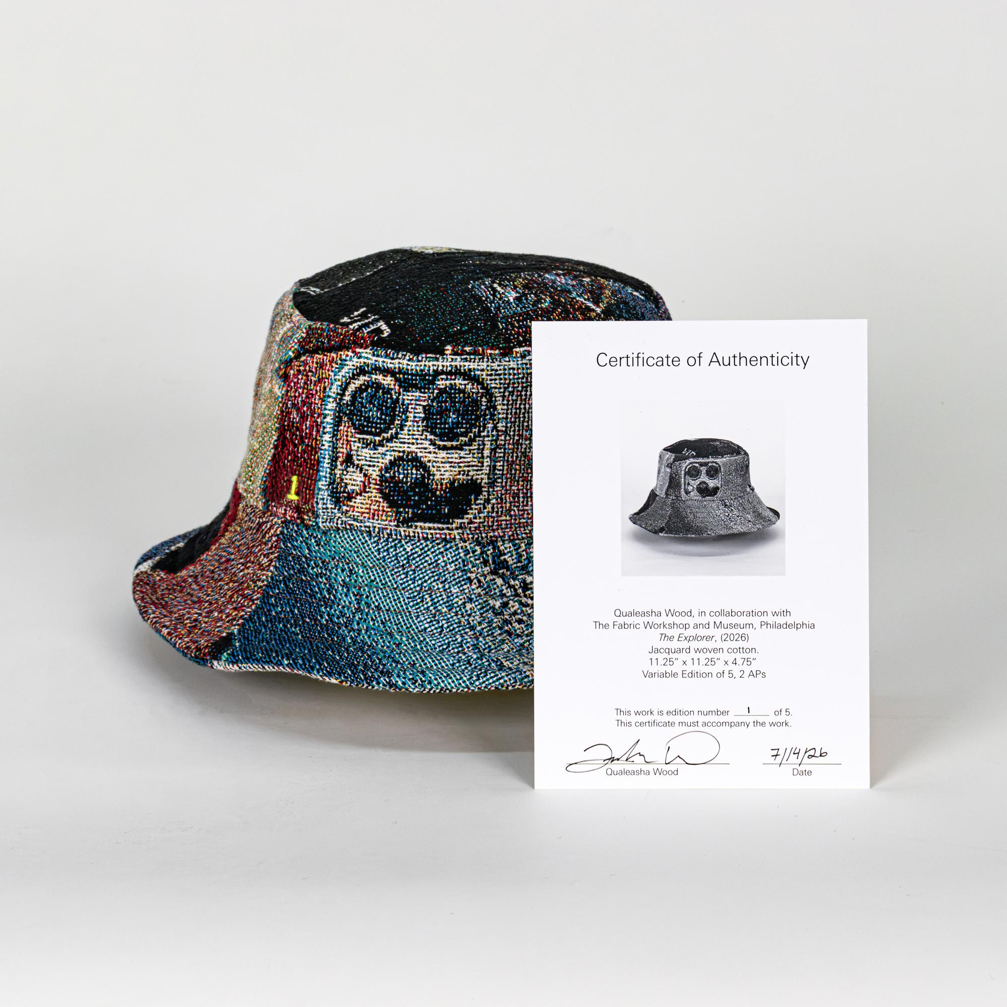 A jacquard-woven bucket hat depicting textures, colors, and symbols of computers and tech. This view specifically features a back camera of an iPhone on the side of the bucket hat. In front of the hat is a certificate of authenticity, signed, numbered, and dated by the artist.