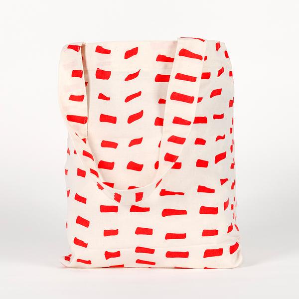 A tall, rectangular tote bag made of white fabric yardage with a printed pattern of short, gestural, brushstroke-like red dashes.