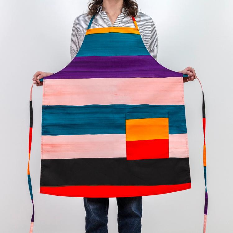 A model wearing a long tie-apron featuring painted horizontal stripes of purple, light pink, teal, red, black, and ochre.