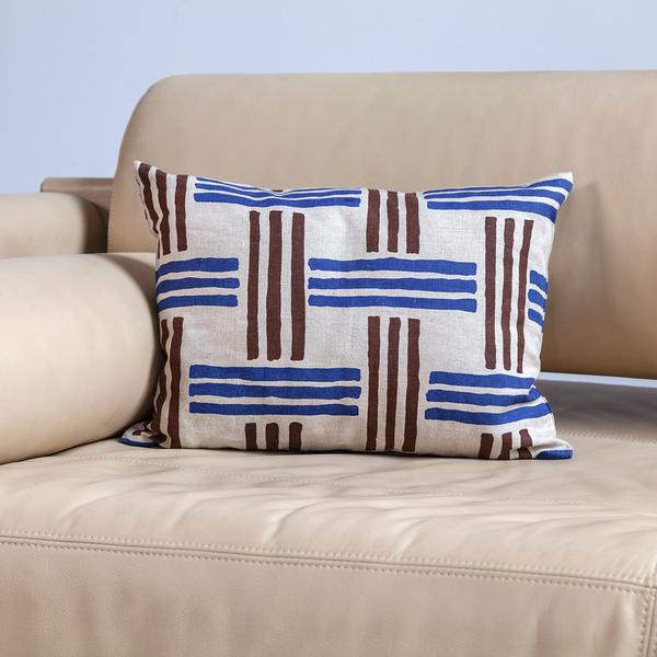 A pillow rests near the corner of a beige couch. The rectangular pillow is made of cream-colored fabric with a minimal weave-like pattern of blue and brown stripes in groups of three.