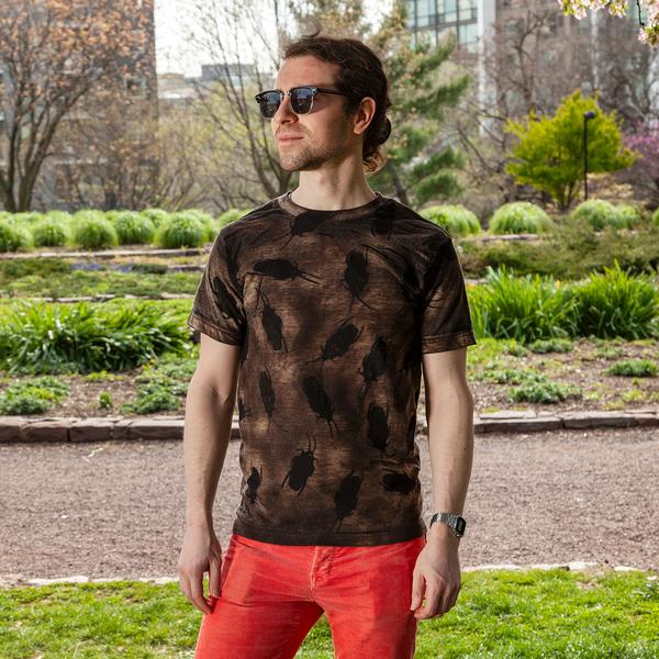 A model wears a mottled brown t-shirt with large black roaches printed across it. He is standing outdoors in a park and wearing sunglasses.