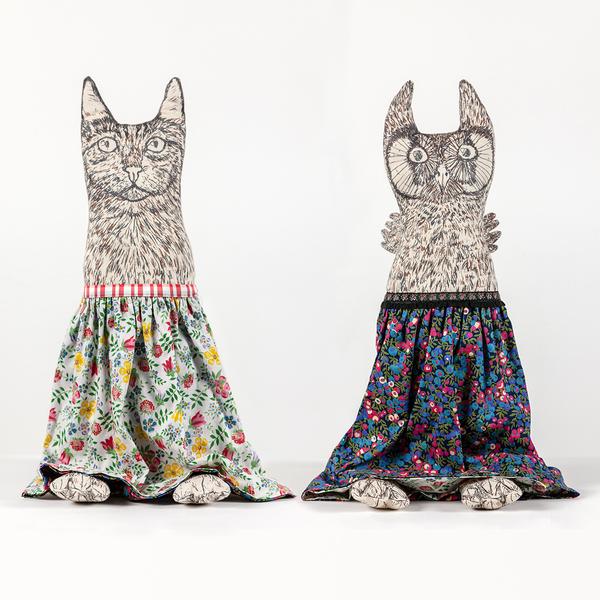 A photograph of two reversible flip dolls side by side. Each doll shows one side of the flip. To the left, the doll shows an image of a cat screenprinted on fabric and wearing an orange and blue open paisley pattern skirt—the cat has little feet peeking out from beneath the skirt's hem. On the right, there is an image of an owl screenprinted on fabric, also wearing a skirt (with a tighter floral pattern in dark blue, light blue, green, red, and white) and with small wings and feet.