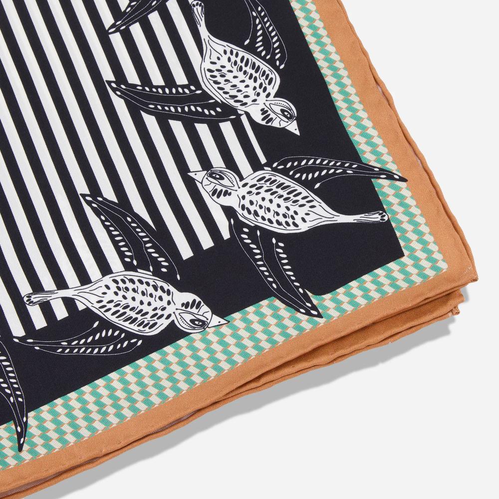 A corner detail of a scarf lined with illustrations of birds. The outer border is tan, followed by a houndstooth green and cream border, followed by a thick black border over which the birds are printed. At its center is a series of black and white stripes.