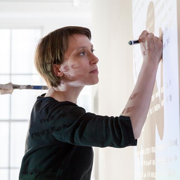 A photograph of a white woman with short brown hair. She is wearing a black shirt with sleeves to her elbow and is drawing on the wall with her right hand, seemingly tracing from a projection.