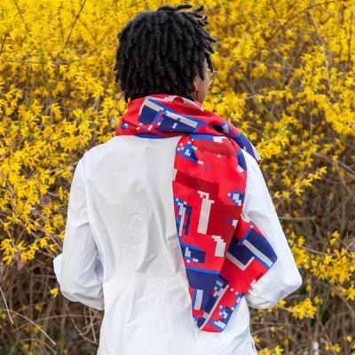 A photo of the back of a young Black woman wearing a bright red silk scarf with blue and white pixelated shapes. The scarf is wrapped around her neck and draped over her shoulder, falling down the back of her white dress shirt. She is looking slightly to her right but we cannot see her face. She is standing in front of a bright yellow forsythia bush.