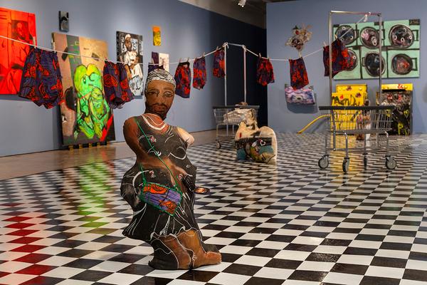 An interior image with black and white checkered floors and blue walls with a variety of paintings and objects hung and leaned against it. In the foreground, a figurative soft sculpture resembling a Black person in a black dress stands upright. Behind them, a clothesline in the background hangs a series of matching blue and red boxers.