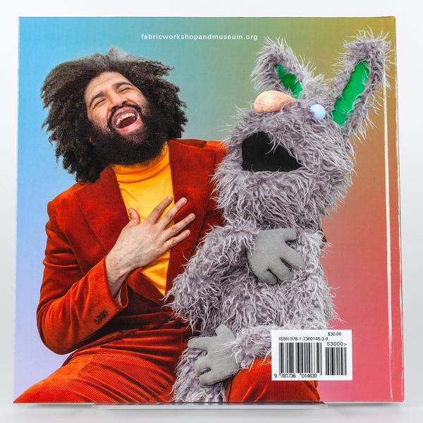 The back cover of a book featuring the artist Jayson Musson and a large rabbit puppet giving a hearty laugh with their hands on their chests. The man is wearing a burgundy corduroy suit with a yellow turtleneck. He has thick dark curly hair that blends in with his beard. The rabbit puppet is grey with stringy fur, a round sky blue eyeball, a pink nose, and green at the center of its tall ears.
