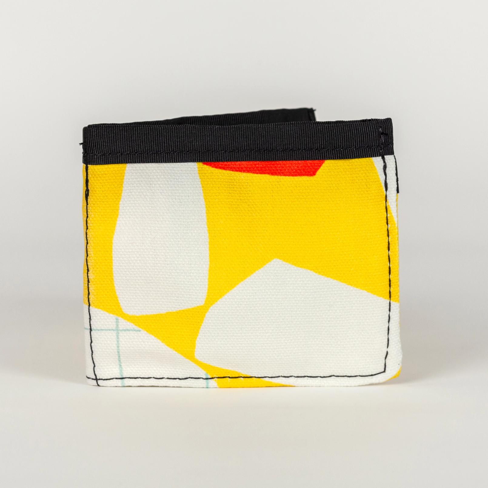 A vibrant yellow wallet folded in half (from being a wide rectangle into a square) with bold abstract shapes (bright orange, white, and white with a light blue grid pattern). There is a solid black seam lining the top of the wallet.