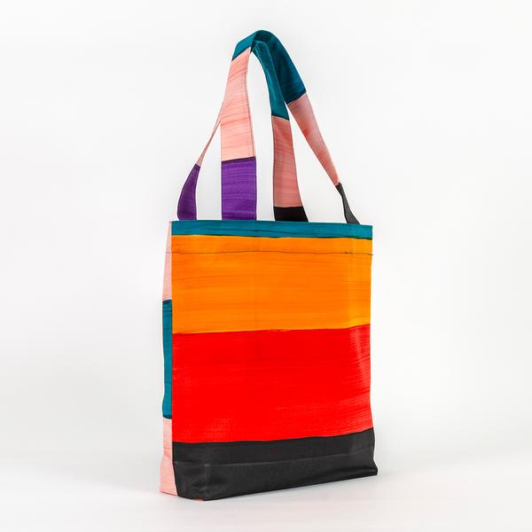 A tote bag taller than it is wide with straps about three-quarters of the bag's height. The bag features a pattern of thick stripes of yellow, red, black, teal, purple, and peach painted with the texture of brush strokes.