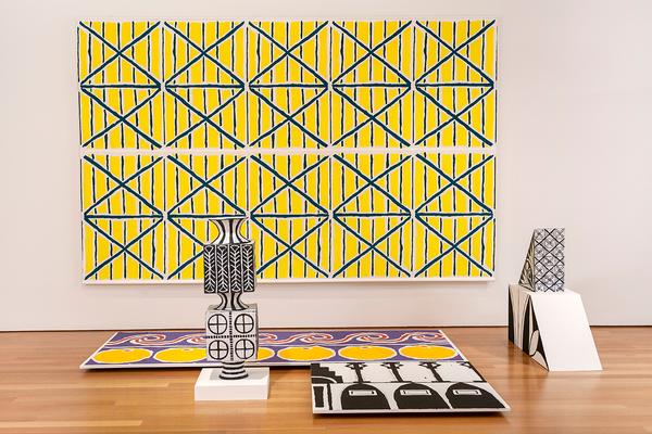 An exhibition view of a large, geometric, abstract repeat pattern in yellow, teal, and white. It is a screenprint and is hanging on the wall like a large painting. In the foreground there are three sculptures decorated with geometric patterns. Two of the sculptures are made of ceramic and are black and white. The third sculpture is a flat panel on the ground with two geometric patterns, one black and white and the other is yellow, purple, and red.