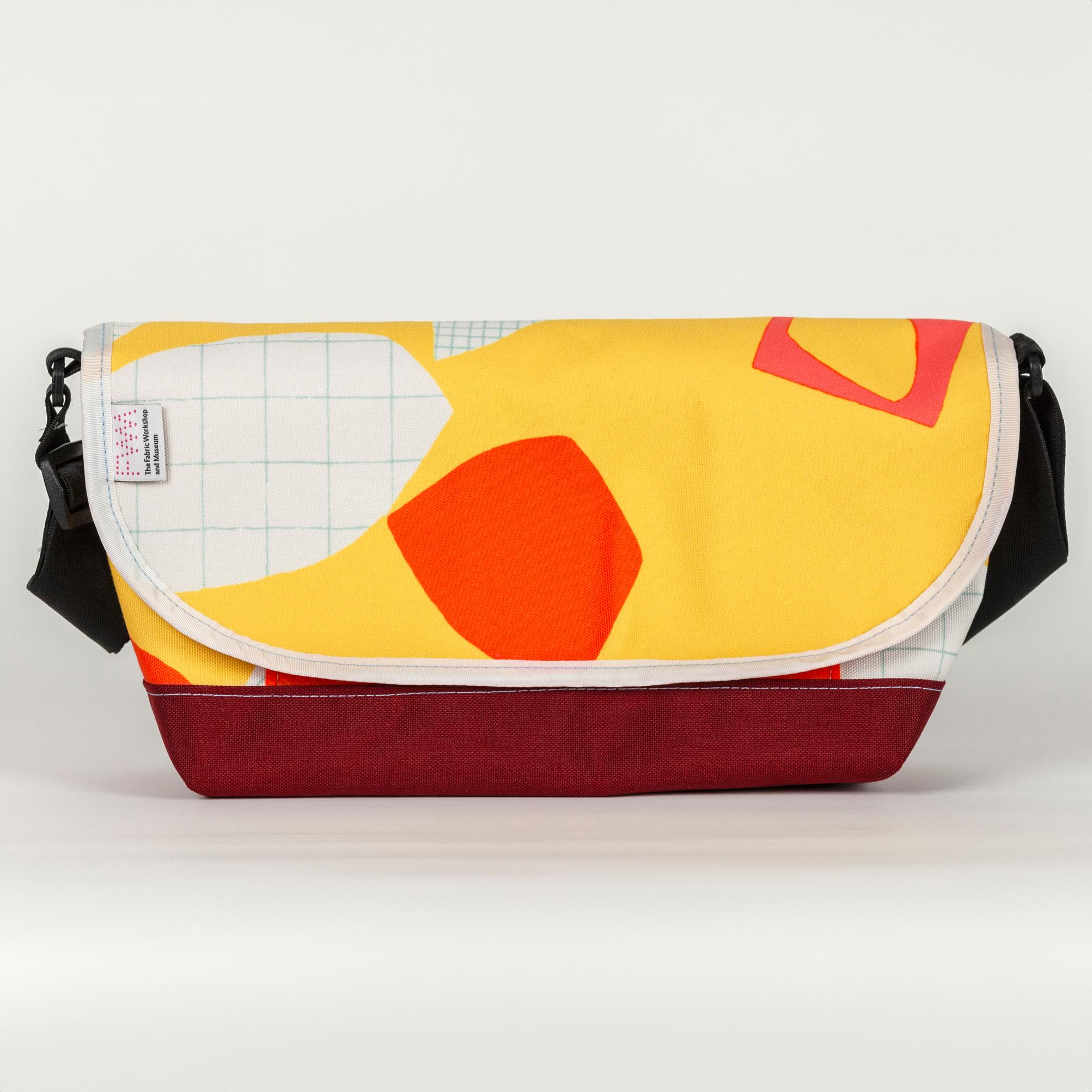 A vibrant yellow bag with bold abstract shapes (bright orange and white with a light blue grid pattern). The bag has a velcro top flap closure with a white seam along the flap. Along the white seam on left side of the bag is a white tag with a pink dotted logo reading "FWM" with "The Fabric Workshop and Museum" written underneath  The bag has a black strap and the base of the bag is solid maroon.