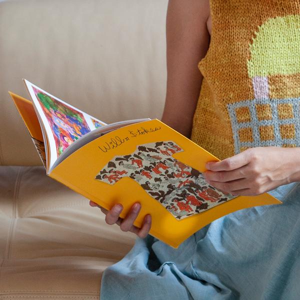 A detail of a person holding a yellow book that reads "Will Stokes". The book cover features the outline of a shirt with many hand-drawn figures in gray, brown, and orange.