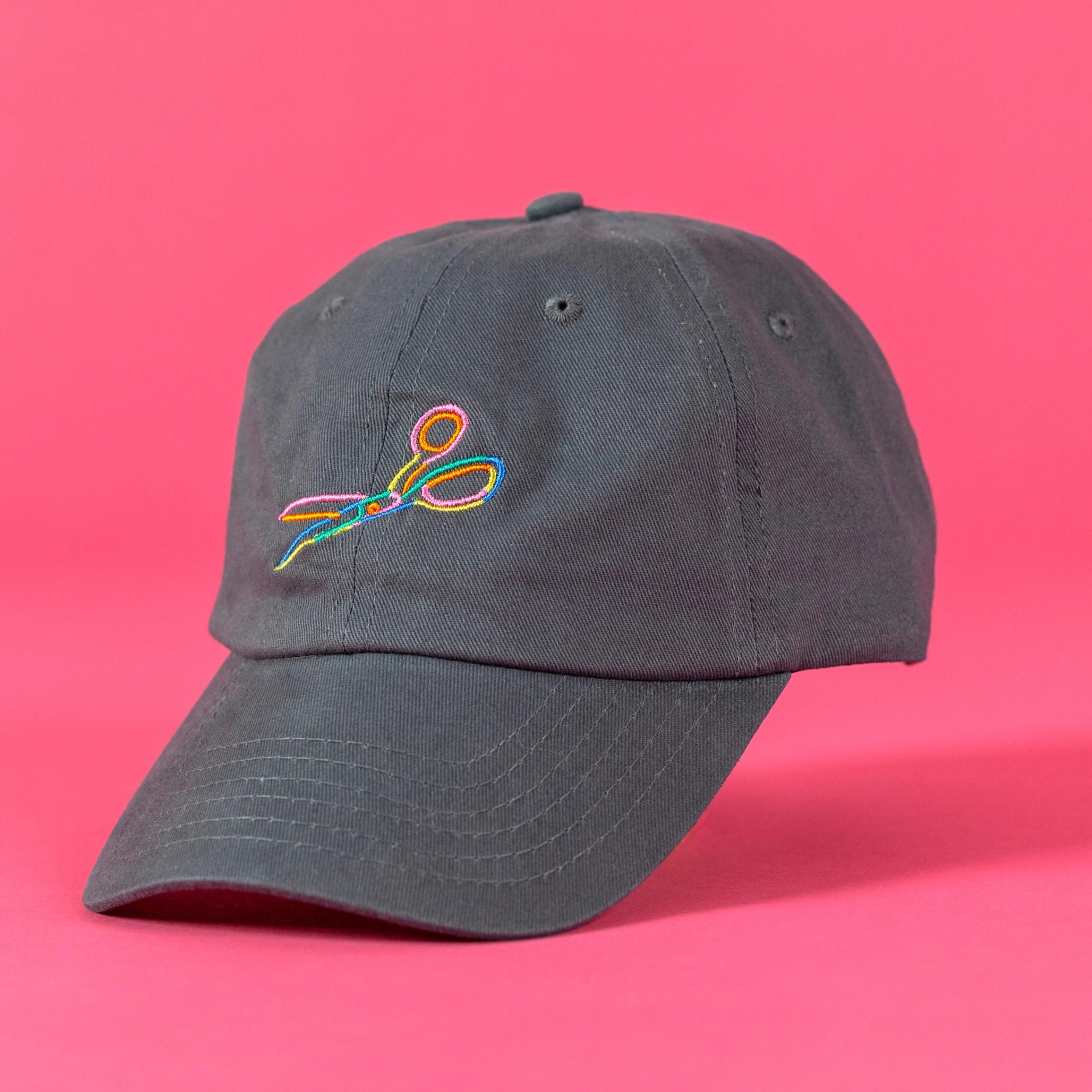A grey baseball cap against a bright pink background. The cap is at a three quarter angle and looks as though it is floating. On the front of it, an illustrated pair of open scissors is embroidered in yellow, orange, blue, green, and pink embroidery threads.  