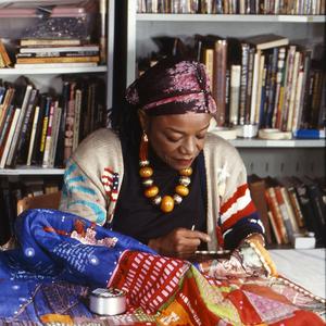 A photograph of Faith Ringgold, a Black woman wearing a headscarf and a beaded necklace, working on a quilt while seated at a table.