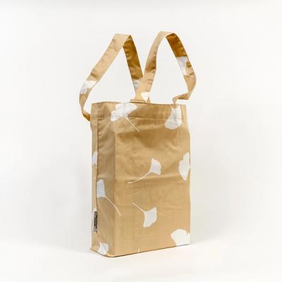 A photo of a tote bag pictured at a slight angle against a white background. The tote's tan fabric features a pattern of white, life-size silhouettes of ginkgo leaves randomly and sporadically spread out around the bag's form.