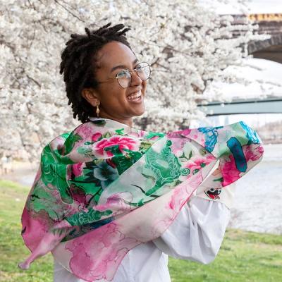 A photograph of a young Black woman smiling as she wraps a scarf around her shoulders while standing on a riverbank in the springtime. The scarf features fluid ink drawings of pink, green, and blue clouds that intersect with drawings of mountains and peonies in an East Asian style.