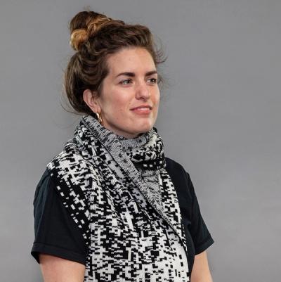 A person wearing a black t-shirt with their hair in a bun against a grey background is wearing the scarf. The scarf is knit in cashmere and features a black and white, pixelated design.
