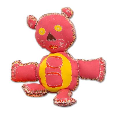 A hand-drawn teddy bear plush doll with a mostly dark pink body, sitting upright. It has yellow eyes, a dark brown wedge shape for a nose, and red lips with smile lines. Its yellow belly features a pink pillar with alternating swaths of red crescents. Its front limbs are significantly larger and longer than its back legs from which it rests. The whole bear figure is outlined in a cream bands with dark marks to suggest stitching.