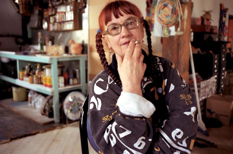 A portrait of an older white woman with red pigtails. She wears glasses and is smoking a cigarette in an artist’s studio.