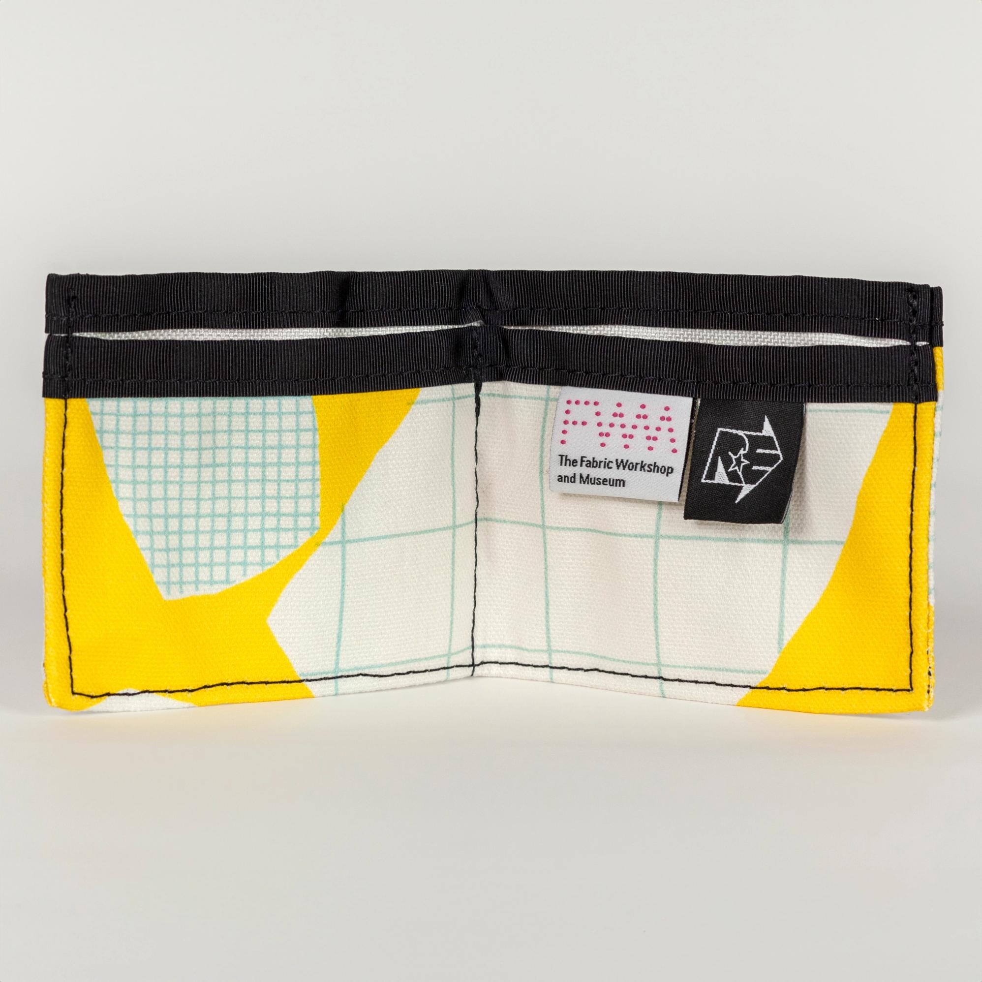 An open vibrant yellow wallets (opens into a wide rectangle with one large pocket in the back and two smaller pockets in front) with bold abstract shapes (white with a light blue grid pattern). There is a solid black seam lining the top of the wallet. There are two tags on the right pocket. The tag on the left is white with a pink dotted logo reading "FWM" with "The Fabric Workshop and Museum" written underneath. The tag on the right is black with an arrow. Inside the arrow are the letters "RE"