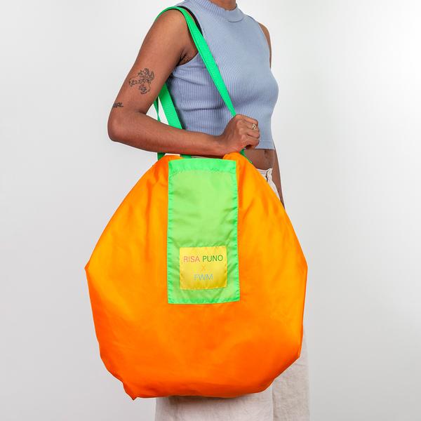A person holds a large nylon bag on their shoulder. The bag is orange with green straps and a yellow tag that reads "Risa Puno x FWM."