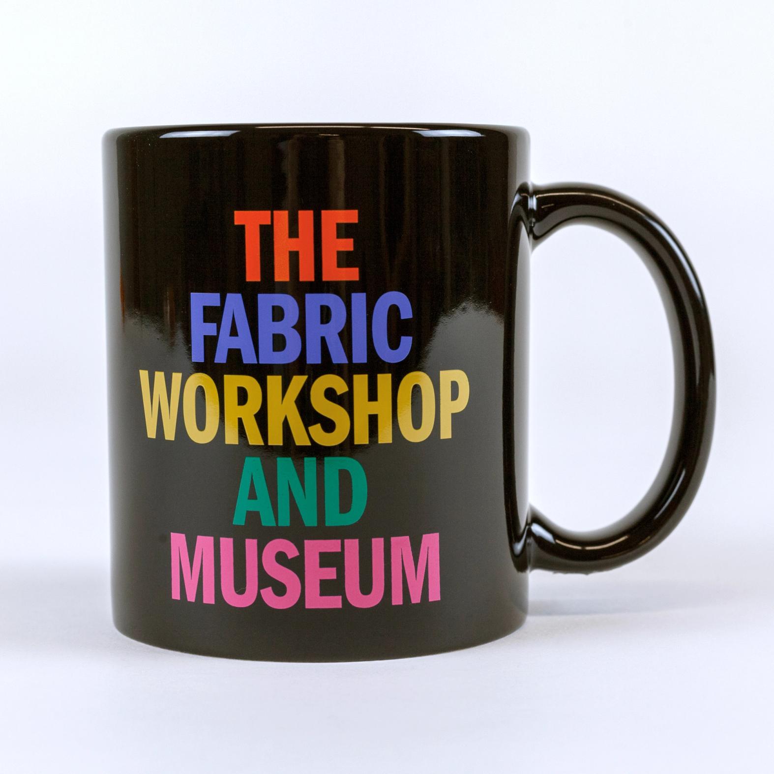 A black colored mug against a white background that reads "THE FABRIC WORKSHOP AND MUSEUM". Each word is stacked and in a different color: orange, blue, golden yellow, green, and pink.
