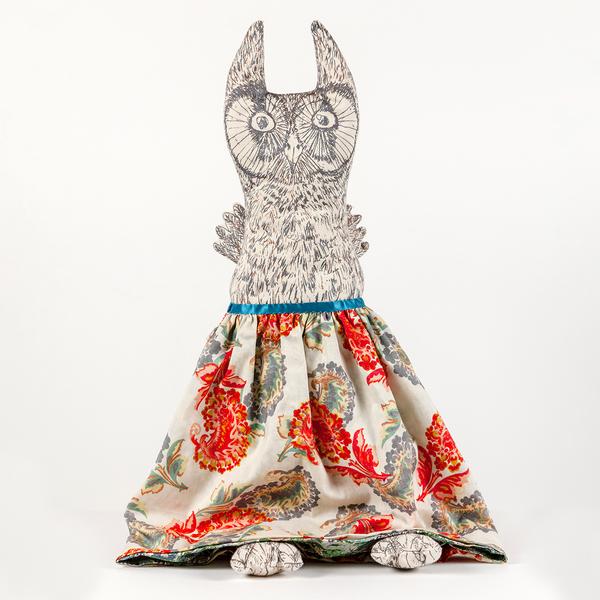 A photograph of a reversible flip doll. In this instance, a plush owl with drawn details stands upright with a skirt of paisleys screenprinted in reds and grays over a cream colored fabric. The owl has little feet peeking out from beneath the skirt's hem.