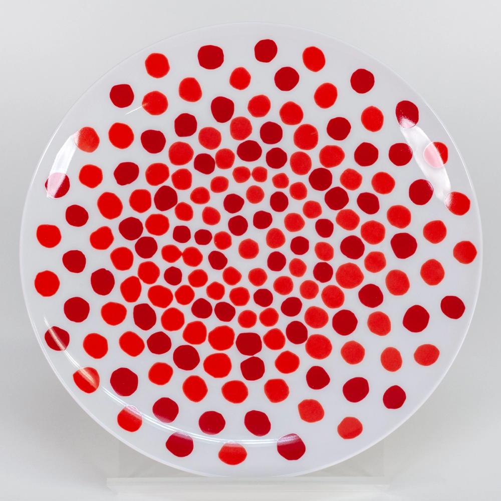 A photograph of a white plate with bold wobbly red dots in wobbly concentric circles. The dots are closer together in the center of the plate and become more spread out towards the edge. The dots do not vary greatly in size.