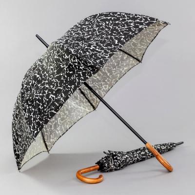 An opened umbrella with a black and white speckled pattern leans diagonally with both part of its shell as well as its wooden handle resting on the same surface. Underneath it is a closed umbrella just like it, lying on the surface and pointing away from the viewer; its wooden handle at the lower center of the image.