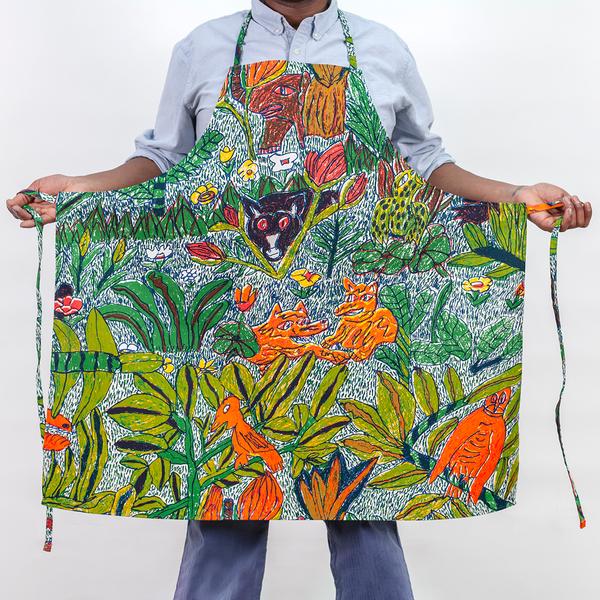 A studio photo of a man wearing a screenprinted apron. The pattern on the apron features a colorful design of hand-drawn leafy foliage, flowers, and wild creatures. The model is holding the apron outwards to show the full width of the apron and the ties.