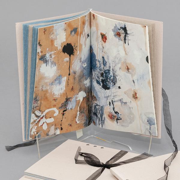 Open paperback book with a textured beige cover, tied with a black ribbon, opened to abstract painted pages in earthy browns, blues, and whites.