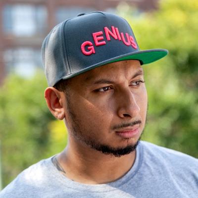 A photograph of a young man wearing a grey cap that reads GENIUS in all caps and in magenta embroidery. He has a thin dark mustache and goatee and is wearing a light gray shirt. The green of trees appears behind him.