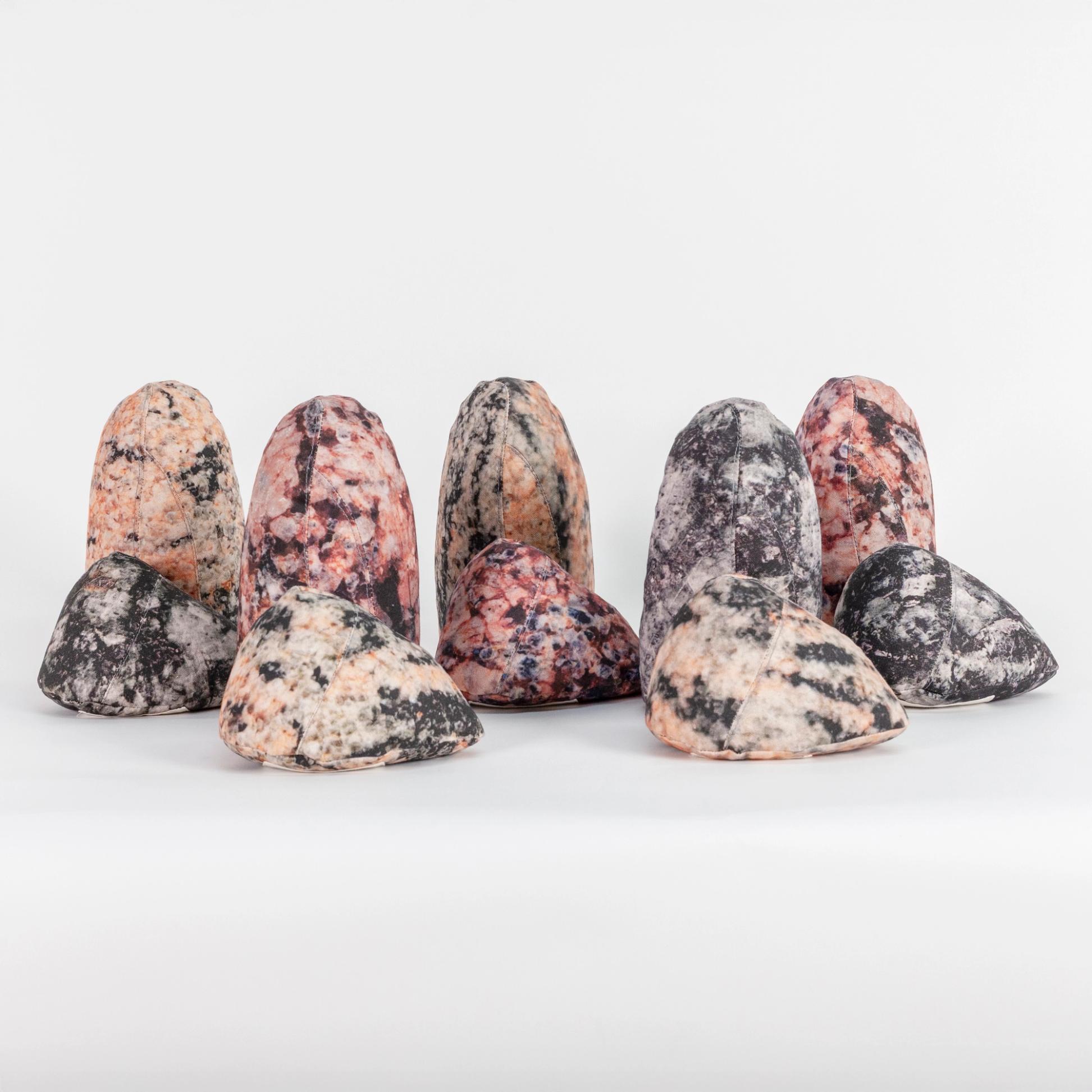 A photograph of a grouping of ten small soft sculptures covered with patterns that resemble polished stone of different hues. Half of the sculptures are short and triangular where the other half are tall like an obelisk with a rounded top.
