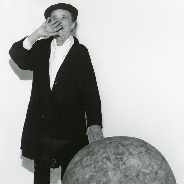 A black and white picture of an older woman, the artist Louise Bourgeois, laughing while waving her right hand over her mouth. She is wearing a black beret and black coat over a white blouse. She is resting her left fingers on a spherical sculpture that rises past her knees.