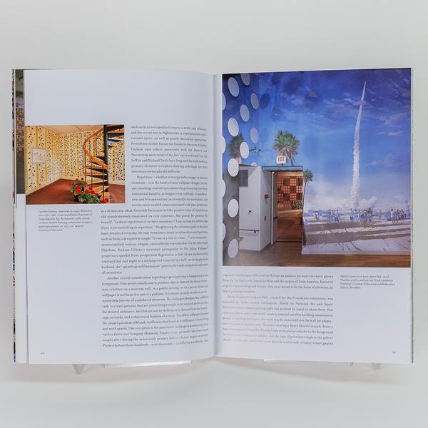 An open book showing a page with text and a small photo of a spiral staircase in a wallpapered room on the left, and a full-page photo on the right of a rocket launch mural with a doorway installation in front of it.