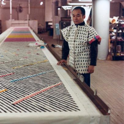 A photograph showing a long table in perspective with black and white and at times colorful striped fabric yardage disappearing as it approaches the far wall. On the right is the artist, Jun Kaneko, a short Japanese man with short hair, observing his work. He is wearing a boxy white tunic with dark long sleeves protruding from underneath. 