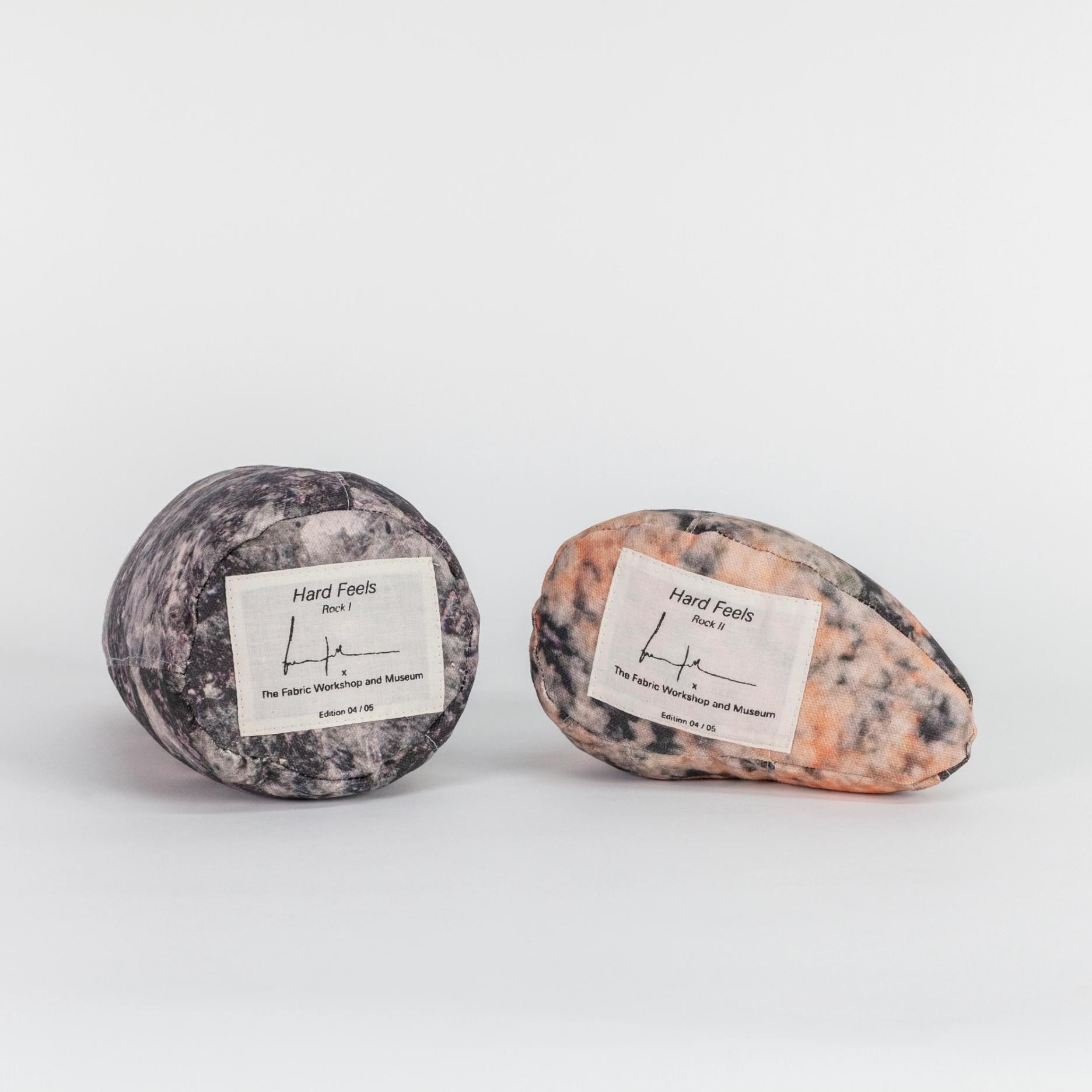 A photograph of the bottom sides of two small soft sculptures covered with patterns that resemble polished stone of different hues. Each one has a label reading the title of the edition (Hard Feels), the artist's signature, and edition number.