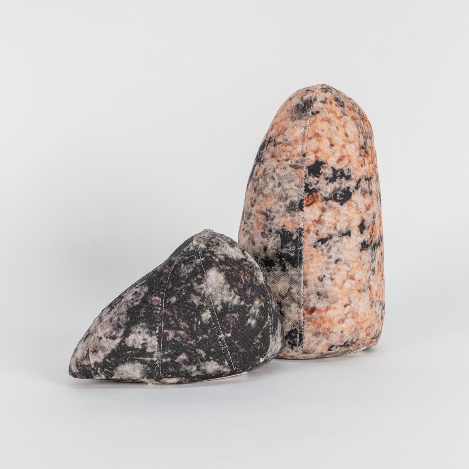 A photograph of two small soft sculptures covered with patterns that resemble polished stone of different hues. One sculpture is short and triangular where the other one is tall like an obelisk with a rounded top.