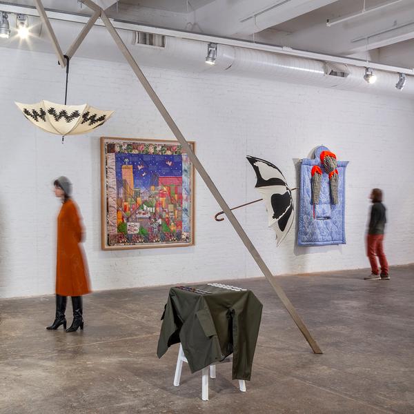 A photo showing two visitors explore an art installation in a gallery. Two umbrellas are installed at the top and further down an armature. Other garments and artworks installed on the floor or wall nearby.