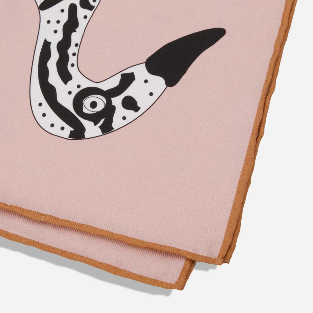 A detail of a folded rose pink silk pocket square with a warm, dark tan bordered seam. There is a glimpse of the head of the printed bold black and white illustration of the Bata bird.