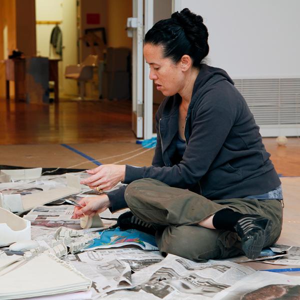 A photograph of a woman sitting cross-legged on the ground on top of a stack of newspapers. She is fixated on the papers and may be rearranging them as her hands are in motion.