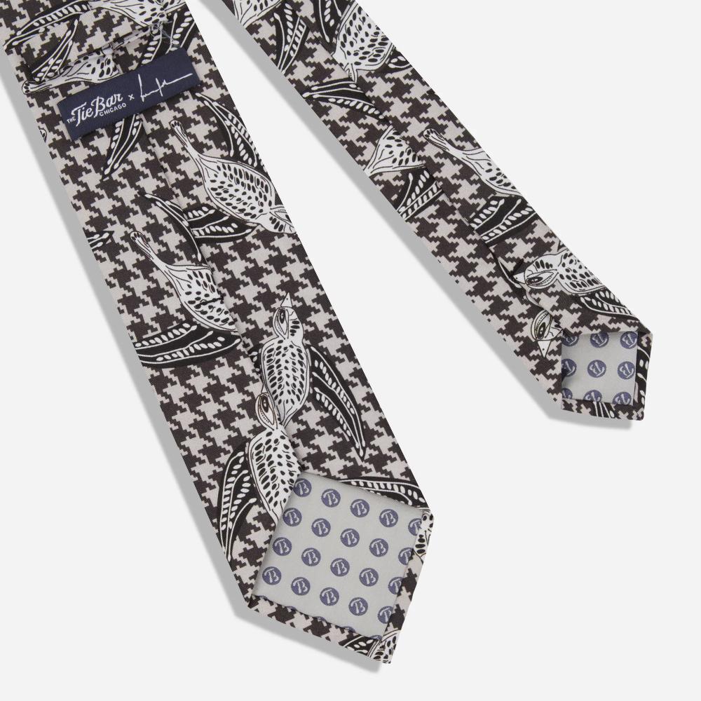 A detail of the back of a tie with a pattern of black and white illustrated Shomoro birds flying in different directions on a light gray and black houndstooth background. There is a label with the artist's signature and "The Tie Bar" logo.