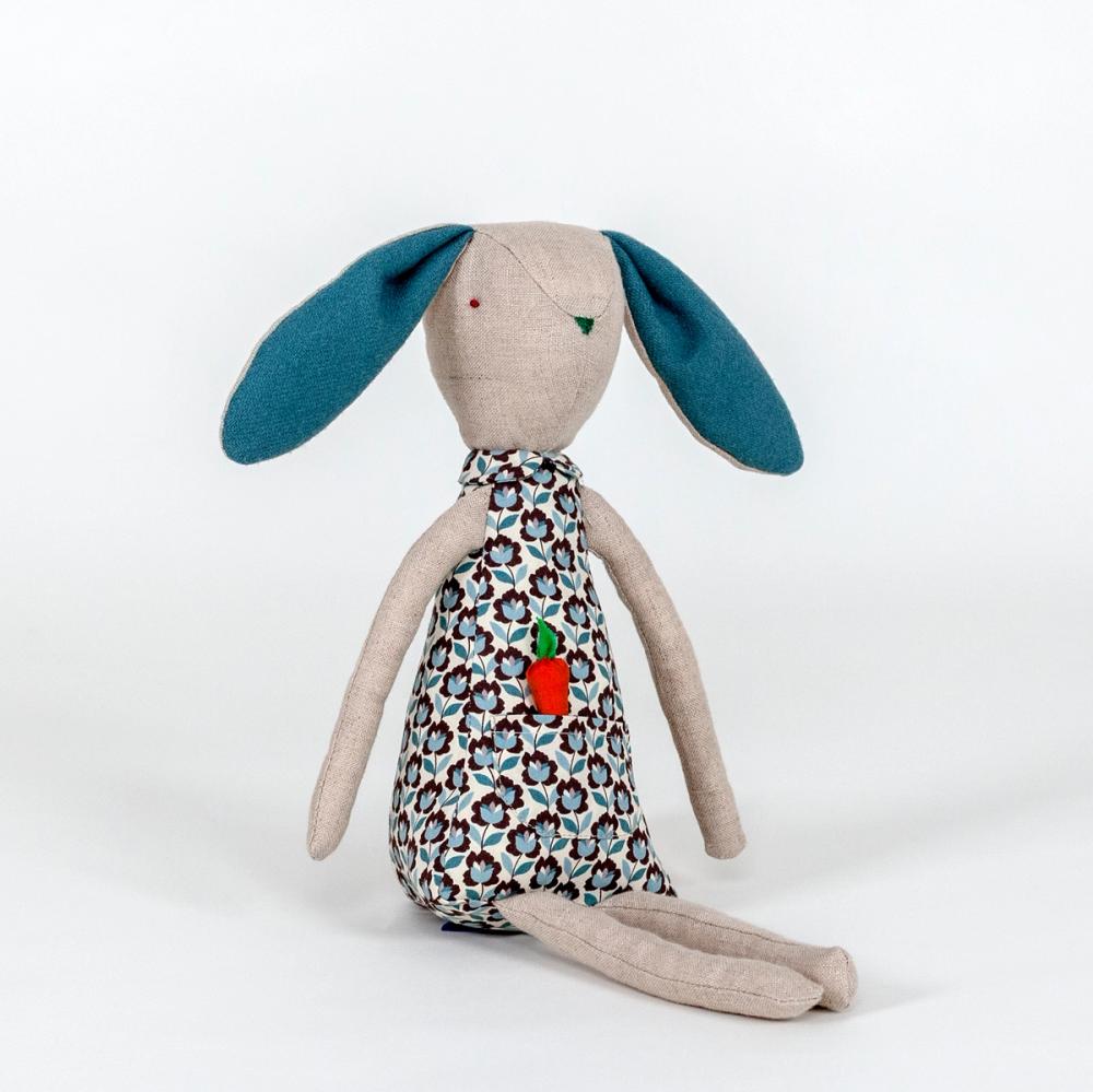 A photograph of a plush rabbit made of light beige woven fabric characterized to have a pointy nose, triangular shaped body, and long thin arms and legs. The body of the rabbit is made of a primarily blue patterned fabric and includes a pocket with a removable plush orange carrot.
