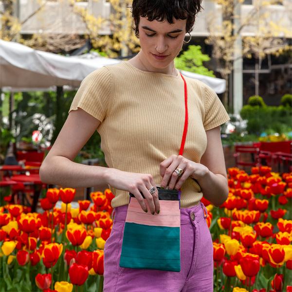 A model begins to pull her phone from a small flat bag on her hip. The bag features stripes of teal, light pink and black, and has a red strap. The person is standing in a park with red and yellow tulips behind her.