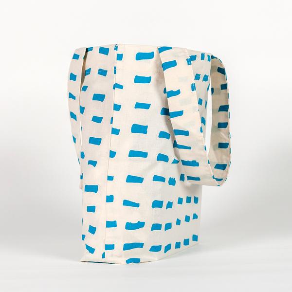 A tall, rectangular tote bag made of white fabric yardage with a printed pattern of short, gestural, brushstroke-like blue dashes.