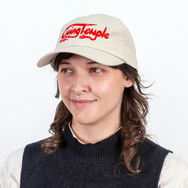 A photograph of a model wearing a tan baseball cap with scripted text reading "Living Temple" embroidered on the front in red thread