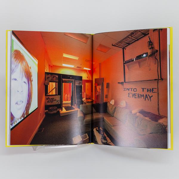 A photograph of an open book featuring a full spread photograph of a room that resembles an office turned living room with orange ambient light and a projection of the face of a young girl on the wall.