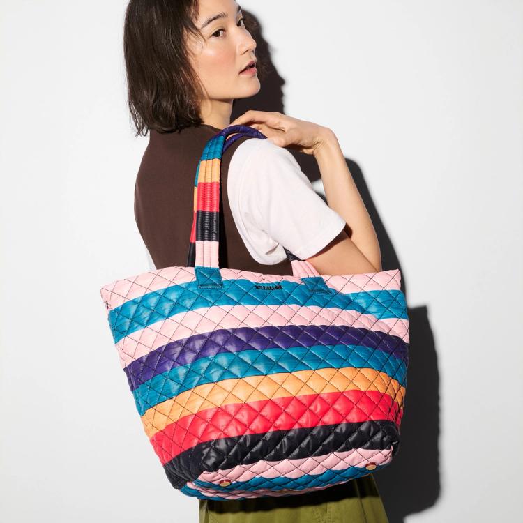 A photograph of a model sporting a fabric quilted tote bag featuring painted horizontal stripes of alternating light pink, purple, blue, ochre, red, and black