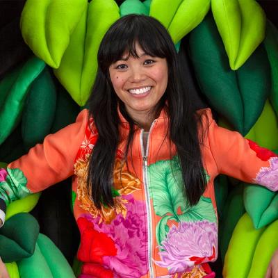 A photograph of the artist Risa Puno, a young woman with black hair, a warm colored blouse and grey jeans. Her recliner-like seat is made of green felted leaves.